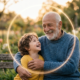Un uomo anziano sorridente abbraccia un bambino piccolo, entrambi seduti su una panchina al tramonto. Un cerchio di luce dorata e soffusa li avvolge, simboleggiando il ciclo della vita e il ritorno alla purezza. Un'immagine calda che rappresenta il legame tra l'alba e il tramonto dell'esistenza.