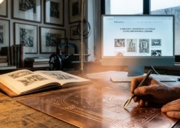 Una mano esperta incide con bulino una lastra di rame in primo piano, simbolo della grafica d'arte. Sullo sfondo, uno schermo digitale acceso mostra il catalogo de 'Il Paladino' con la scritta olografica 'Resistenza culturale. Oltre l'informatica, l'Essere'. Un ponte tra tradizione incisoria e mondo digitale.
